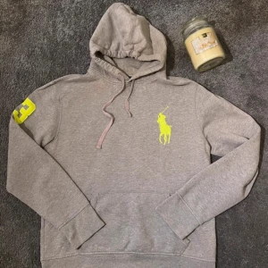 Grå hoodie från Polo Ralph Lauren med neongula detaljer - Säljer en grå hoodie från Polo Ralph Lauren med stor neongul logga på bröstet och siffran 3 på ärmen. Tröjan har huva med dragsko och magficka. Perfekt för en sportig och avslappnad stil.