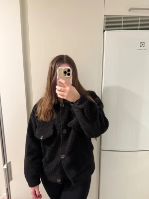 Svart teddyjacka med knappar - Mysig svart teddyjacka med stora knappar framtill och två fejk fickor på bröstet. Jackan har en loose passform och är långärmad med fluffigt, mjukt material som ger en cool och chill vibe. Perfekt för dig som gillar oversized och vill ha något riktigt skönt.