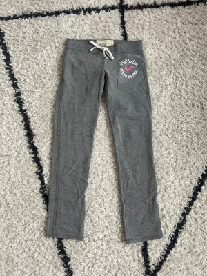 Grå mjukisbyxor Hollister 🩶 - Grå mjukisbyxor från Hollister med broderad logga och texten 'Ocean Patrol' i vitt och rosa på vänster höft. Byxorna har normal passform, raka ben, hyfsat låg midja och vit snörning i midjan 🩶