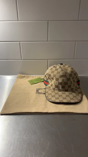 Gucci beige keps med GG-mönster - Snygg beige keps från Gucci med klassiskt GG-monogram över hela kepsen och gröna/röda ränder på sidorna. Justerbar rem baktill och detaljer i brunt. Materialet är bomull och syntet, perfekt för dig som vill ha en lyxig streetstyle-look.