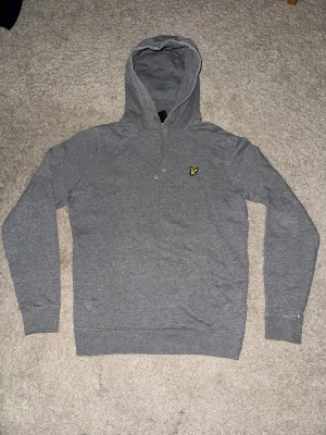 Grå hoodie från Lyle & Scott - Snygg grå hoodie från Lyle & Scott med klassisk gul fågel-logga på bröstet. Tröjan har huva med snörning, magficka och ribbade muddar vid ärmar och nederkant. Perfekt för chill dagar och enkel att matcha med jeans eller joggers.