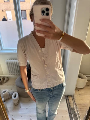 Ljusbeige kortärmad blus med knappar - Säljer en ljusbeige blus med korta ärmar och ribbade detaljer vid ärmslut, nederkant och längs knappslån. Blusen har v-ringning, två små fickor framtill och stora vita knappar hela vägen. Perfekt till jeans eller kjol för en chill och fräsch look.