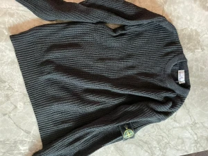 Mörkgrå stickad tröja Stone Island - Säljer en mörkgrå stickad tröja från Stone Island med klassisk rund hals och ribbad struktur. Tröjan har lång ärm och den ikoniska Stone Island-loggan på vänster ärm. Perfekt för dig som gillar stilrena och coola plagg.