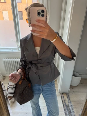 Grå omlottkavaj med bälte - Snygg grå kavaj med omlottmodell och knytbälte i midjan. Kavajen har breda slag, knappar framtill och trekvartsärmar. Materialet har en lätt satinlik finish som ger en cool vibe. Perfekt att styla med jeans för en trendig look.