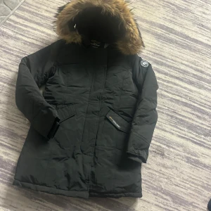 Svart parkasjacka med päls från Canada goose black label  - Svart varm Stillig parkasjacka från Canada  goose black label med huva och snygg äkta racon pälsdetalj i brunt. Jackan har långa ärmar, två stora fickor framtill och ett märke på ena ärmen. Perfekt för kalla dagar med sin varma och långa design.