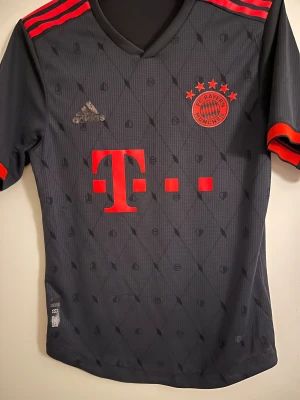 FC Bayern München svart matchtröja - Snygg FC Bayern München fotbollströja från Adidas i svart med röda detaljer och logga. Tröjan har korta ärmar, V-ringning och ett diskret rutmönster. Materialet är lätt och andas, perfekt för träning eller match. Ikoniska Adidas-ränder på axlarna och stor T-Mobile sponsorlogga på bröstet.