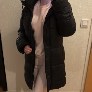 Svart lång pufferjacka med huva - Säljer en svart, lång pufferjacka med huva och snörning. Jackan har quiltad design, dragkedja framtill och stora, mysiga ärmar. Perfekt för kalla dagar när du vill hålla dig varm och ändå se chill ut.