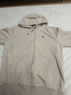 Beige hoodie från Polo Ralph Lauren - Snygg beige hoodie från Polo Ralph Lauren i 100% bomull. Tröjan har huva med snörning, dragkedja framtill och två stora fickor. Klassisk mörkblå logga broderad på bröstet. Storlek M men är nog lite större än det. Lägg bud.
