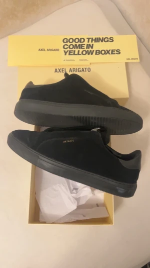 Svarta sneakers från Axel Arigato - Snygga svarta sneakers från Axel Arigato med minimalistisk design och diskret guldfärgad logga på sidan. Skorna har en platt sula och är tillverkade i mjuk mocka med detaljer i skinn. Perfekta för dig som gillar stilrena och trendiga skor.
