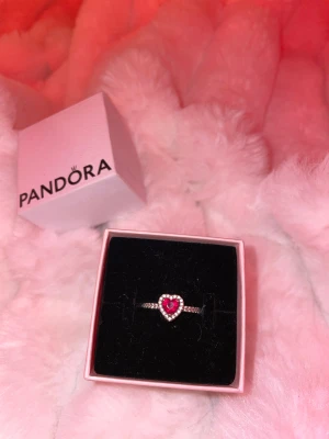 Pandora silverring med hjärta - Säljer då min ring som inte kommer komma till användning något mer, det är en snygg silverring från Pandora med ett rött hjärta i mitten, omringat av små glittrande stenar. Ringen har en tunn, elegant form och kommer i original ask. Perfekt till allt