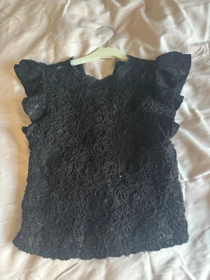 Svart spetsblus med volangärmar - Supersnygg svart blus i spets med blommigt mönster och volangdetaljer på ärmarna. Blusen är ärmlös med lätt v-ringning och har en romantisk vibe. Perfekt att styla med jeans eller kjol för en trendig look.