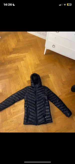 Svart McKinley pufferjacka med huva - Svart pufferjacka från McKinley med huva och dragkedja framtill. Jackan har quiltad design och två sidofickor med dragkedja. Perfekt för kyliga dagar och har en clean look som funkar till det mesta. Materialet är syntet och jackan är långärmad.