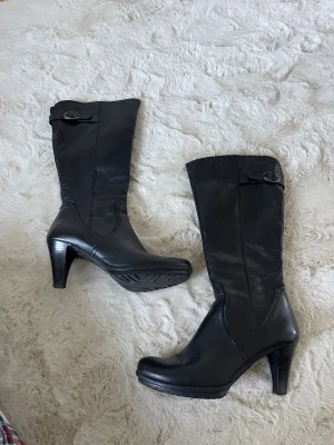 Svarta stövlar med klack från Tamaris - Snygga svarta stövlar från Tamaris i skinn med rund tå och hög klack. Stövlarna har en dekorativ rem med spänne upptill och en plattformsula som ger extra komfort. Köpta på Sellpy men aldrig använda😚💕😇