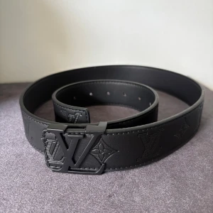 Svart Louis Vuitton skärp i skinn - Säljer ett svart Louis Vuitton bälte i skinn med präglade LV-loggor och monogram längs hela remmen. Bältet har en stilren, matt svart LV-spänne och är tillverkat i Spanien. Perfekt för dig som vill ha en lyxig och snygg accessoar med ikonisk design.