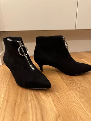 Svarta stövletter med spetsig tå - Snygga svarta stövletter i mocka med spetsig tå och låg klack. Framtill finns en grov silverfärgad dragkedja med stor ring som detalj. Skorna har en elegant siluett och passar perfekt till en trendig look.