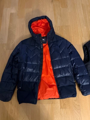 Mörkblå pufferjacka från Mountain Works - Säljer en snygg mörkblå pufferjacka från Mountain Works med orange foder. Jackan har huva, dragkedja och quiltad design. Perfekt för kalla dagar och har en cool kontrast mellan det mörka yttertyget och det orangea fodret.