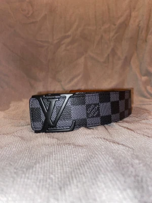 Louis Vuitton rutig svart bälte - Säljer ett Louis Vuitton bälte med ikoniskt rutigt mönster i svart och grått. Bältet har en stor LV-logga i svart metall som spänne och är tillverkat i skinn med beige insida. Perfekt accessoar för att lyfta din outfit.