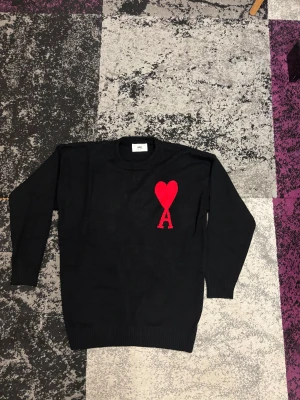 Svart stickad tröja från Ami Paris - Svart stickad tröja från Ami Paris med lång ärm och rund hals. På bröstet finns ett stort rött hjärta med bokstaven A, vilket ger en unik och trendig look. Tröjan har ribbade muddar vid ärmslut och nederkant för extra komfort.