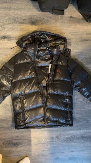 Svart pufferjacka från Polo Ralph Lauren - Svart pufferjacka från Polo Ralph Lauren med glansig finish och röd logga på bröstet. Jackan har huva, dragkedja framtill och två sidofickor med dragkedja. Perfekt för kalla dagar och har en modern, oversized look.