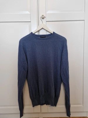 Blå stickad tröja från Best Company - Snygg blå stickad tröja från Best Company med rund hals och ribbade muddar. Tröjan har långärmade ärmar och gråa förstärkningar på armbågarna för en cool detalj. Perfekt för dig som gillar enkel och stilren design.