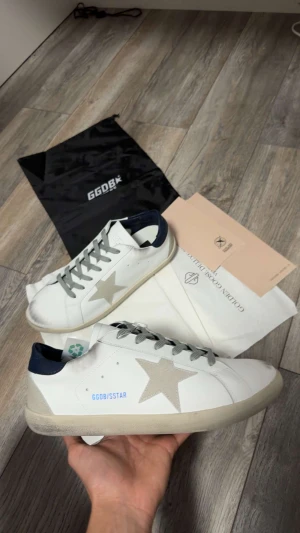 Golden Goose vita sneakers med stjärna - Golden Goose sneakers i vitt skinn med grå mockadetaljer och en stor stjärna på sidan. Skorna har grå snörning, mörkblå häl i skinn och platt sula. Snygg kontrast mellan material och färger, perfekt för dig som gillar streetstyle med exklusiv känsla.