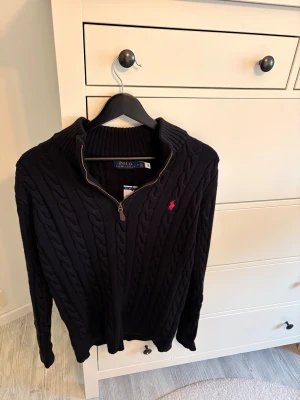  stickad tröja Polo Ralph Lauren - Säljer en svart stickad tröja från Polo Ralph Lauren i storlek M. Tröjan har kabelstickat mönster, half zip-dragkedja och långärmad design. Klassisk röd logobrodering på bröstet och ribbade muddar vid ärmslut och nederkant. Perfekt för kyliga dagar.