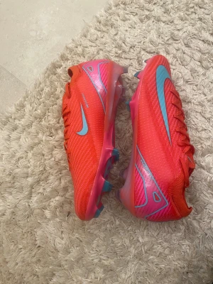 Nike Mercurial röda fotbollsskor - Säljer ett par Nike Mercurial fotbollsskor i en riktigt snygg röd färg med ljusblå swoosh och detaljer. Skorna har snörning, platt sula med dobbar och är tillverkade i syntetmaterial för bästa grepp och komfort på planen. Perfekt för dig som vill sticka ut på matchen.
