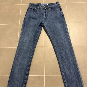 Blå jeans från Jacob Cohen, storlek 32 - Snygga blå jeans från Jacob Cohen i klassisk straight fit med fem fickor och dragkedja. Jeansen har en mörkblå tvätt, silverfärgad knapp och diskreta sömmar. Materialet är mjukt jeans och insidan har randigt tyg vid linningen. Perfekt för dig som gillar stilrena och tidlösa jeans.