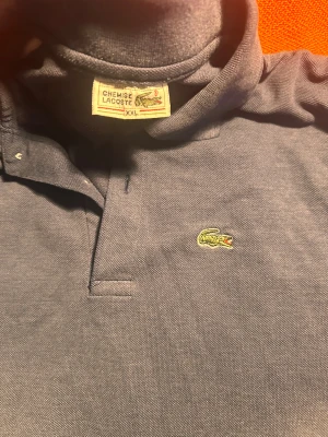 Marinblå pikétröja Lacoste XXL - Snygg marinblå pikétröja från Lacoste i storlek XXL. Klassisk krage, korta ärmar och den ikoniska krokodilloggan broderad på bröstet. Tröjan är gjord i mjuk bomull som känns skön mot huden och har en stilren look som funkar till många outfits.