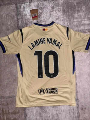 Beige FC Barcelona Lamine Yamal tröja - Säljer en beige FC Barcelona fotbollströja med Lamine Yamal och nummer 10 på ryggen. Tröjan har svarta och blå detaljer vid krage och ärmslut, samt tryck från Spotify och UNHCR. Tillverkad i lätt Dri-Fit material, perfekt för match eller träning.