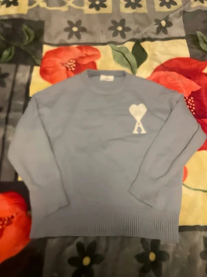 Blå stickad tröja från Ami Paris - Blå stickad tröja från Ami Paris med ett vitt hjärta och bokstaven A broderat på bröstet. Tröjan har rund hals, långa ärmar och breda ribbade muddar vid ärmslut och nederkant. Perfekt för dig som gillar stilrena detaljer.