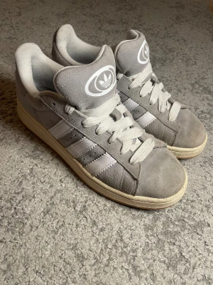 Grå adidas Campus sneakers i mocka - Säljer ett par grå adidas Campus sneakers med klassiska vita ränder på sidan och stor vit logga på plösen. Skorna har rund tå, platt sula och är tillverkade i mjuk mocka med vita snören. Perfekt för dig som gillar retrostil och streetwear. använda fåtal gånger som nya.