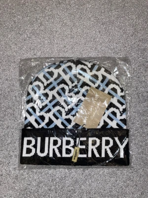 Burberry stickad mössa med logga - Snygg stickad mössa från Burberry med stor vit logga framtill. Mössan har ett grafiskt mönster i svart, vitt och ljusblått, samt bred uppvikt kant. Perfekt för dig som vill ha en cool och lyxig accessoar till vintern.
