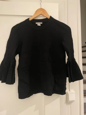 Svart tröja med volangärm från H&M - Svart tröja från H&M i storlek XS med rund hals och långa ärmar som avslutas med breda volanger. Tröjan har en stilren look och är tillverkad i ett mjukt material som känns skönt mot huden. Perfekt för dig som gillar detaljer och vill ha något som sticker ut.