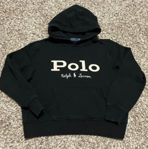 Svart hoodie från Polo Ralph Lauren - Svart hoodie från Polo Ralph Lauren med stor vit logotyp på bröstet. Tröjan har huva, magficka och ribbade muddar vid ärmslut och nederkant. Perfekt för chill dagar och streetstyle. Klassisk och enkel design som funkar till det mesta.