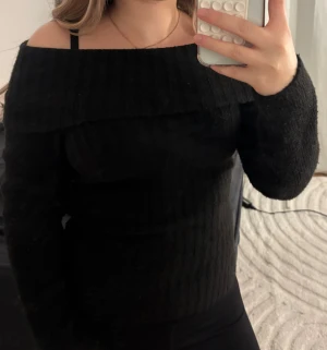 Svart offshoulder stickad tröja - Snygg svart stickad tröja med offshoulder-design och ribbad struktur. Tröjan har lång ärm och sitter tight mot kroppen, perfekt för dig som gillar en stilren look med lite edge. Passar dig som vill ha något enkelt men ändå trendigt.