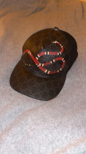 Gucci keps med orm och GG-mönster - Snygg svart Gucci keps med klassiskt GG-mönster och en fet röd och vit orm på framsidan. Kepsen har mesh på sidorna och justerbar rem bak. Tillverkad i Italien, materialet är polyester och bomull. Perfekt för dig som vill sticka ut med designer vibes.