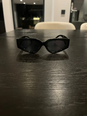 Svarta solglasögon från Chanel - Snygga svarta solglasögon från Chanel med chunky bågar och ikoniska CC-loggan på sidorna. Glasögonen har en modern, kantig form och är tillverkade i blankt syntetmaterial. Perfekt accessoar för att lyfta din stil.