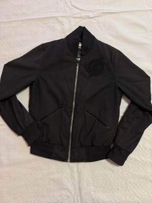 Svart bomberjacka från G-Star RAW - Snygg svart bomberjacka från G-Star RAW med coolt RAW-märke på bröstet och dragkedja framtill. Jackan har ribbade muddar, två fickor med dragkedja och en schysst urban look. Perfekt för dig som vill ha en stilren och trendig jacka till vardags.