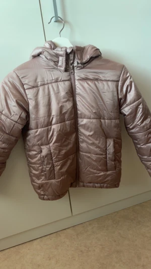 Beige pufferjacka från Lindex Kids - Säljer en beige pufferjacka från Lindex Kids i storlek 134. Jackan har huva, två fickor framtill och dragkedja hela vägen upp. Perfekt för kyliga dagar och har en snygg, enkel design som passar till det mesta.
