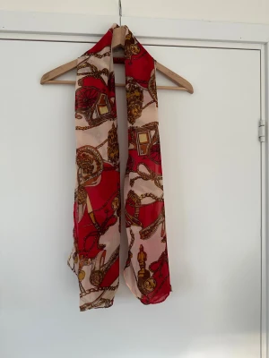 Röd och beige mönstrad halsduk / scarf - Snygg och tunn halsduk / scarf / sjal i rött och beige med detaljerat mönster av kedjor, vagnar och accessoarer i guld och brunt. Materialet är lätt och luftigt, perfekt att slänga över axlarna för en cool touch. Mönstret ger en lyxig vibe och färgerna poppar verkligen.
