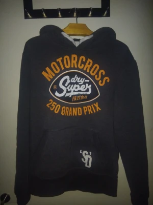 Superdry hoodie limited edition  - Säljer en mörkgrå hoodie från Superdry, Limited Edition Vintage Goods. Stor tryckt logga i orange och vitt på bröstet, med texten 'MOTORCROSS 250 GRAND PRIX'. Klassisk känguruficka och huva. Perfekt för dig som gillar streetwear och snygga detaljer.