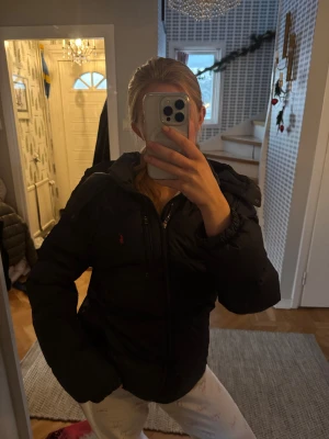  Ralph Lauren jacka  - Pufferjacka från Polo Ralph Lauren med huva och röd logga på bröstet. Jackan har flera dragkedjor, quiltad design och elastiska muddar vid ärmslut. Perfekt för kalla dagar och har en clean, sportig look som funkar till jeans eller mjukisar.