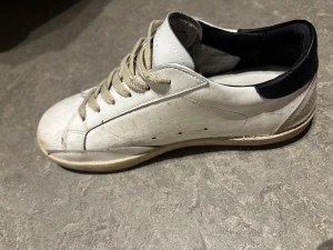 Golden goose - Säljer ett par vita Golden Goose Superstar sneakers med beige stjärndetalj på sidan, svart häl och beige mockadetaljer. Skorna har snörning och platt sula, samt en rund tå. Materialet är skinn och mocka, med en cool vintage-look och kontrastfärgade detaljer.