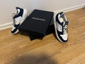 Valentino bounce trainers (säljs inte längre) - Snygga Valentino sneakers i marinblått och vitt med tydlig logga på sidan och bak på sulan. Skorna har snörning, rund tå och platt sula. Materialet är en mix av skinn och mocka som ger en lyxig känsla. Perfekta för dig som gillar exklusiva märken och stilren design. Nypris 5500