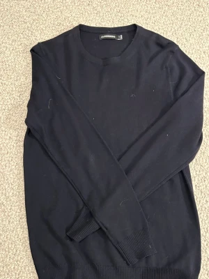 Mörkblå J.Lindeberg Merino Tröja - Snygg mörkblå stickad tröja från J.Lindeberg i storlek M. Tröjan har rund hals, långa ärmar och diskret logga på bröstet. Ribbstickade muddar vid ärmslut och nederkant ger en clean look. Perfekt för dig som gillar stilrena och klassiska plagg.