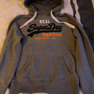  Superdry hoodie  - Säljer en grå hoodie från Superdry med stort svart och vitt tryck på bröstet samt orange text. Tröjan har huva med vita snören, magficka och ribbade muddar. Perfekt för chill dagar och streetstyle. Materialet är mjuk bomull och polyesterblandning.