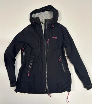 Svart skaljacka från Bergans, L - Svart skaljacka från Bergans med huva och rosa detaljer. Jackan har flera dragkedjor framtill samt rosa logga på ärmen. Justerbar nederkant och ärmslut med kardborre. Lite smutsig. 
