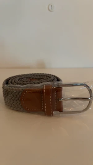 Grått flätat bälte med skinn-detalj - Snyggt grått flätat bälte med bruna skinn-detaljer och silvrigt spänne. Bältet har en bred, flätad struktur som ger en cool och modern look. Perfekt för att lyfta din outfit med en stilren accessoar. Passar till jeans, chinos eller shorts.