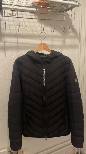 Svart pufferjacka från EA7 Emporio Armani - Svart pufferjacka från EA7 Emporio Armani med huva och snygga diagonala quiltade sömmar. Jackan har dragkedja framtill och EA7-logga på ärmen. Perfekt för kalla dagar och har en modern, sportig vibe.